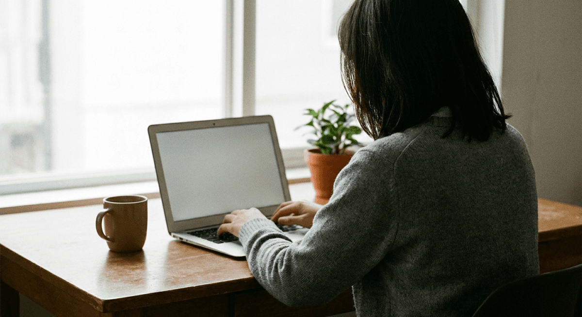 person in a grey sweater typing at a laptop on a wooden desk, seen from behind